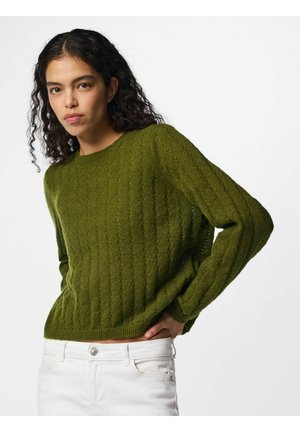 Jeune femme aux cheveux bouclés foncés portant un pull vert texturé et un pantalon blanc, posant avec une main sur la hanche sur fond blanc.