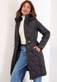 Langer schwarzer gesteppter Daunenmantel mit Kapuze, tailliertem Bund, detaillierter Nahtverarbeitung, seitlichen Taschen und einem Frontreißverschluss. Tragen über einem Rollkragenpullover und Jeans.