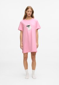 Femeie îmbrăcată cu o rochie supradimensionată roz, cu un grafic al unei pisici albe purtând ochelari de soare negri, stând desculță cu șosete albe pe un fundal alb.