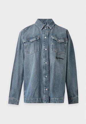 Femme aux longs cheveux blonds portant une veste en jean bleue délavée et oversize ainsi qu’un jean à jambes larges, debout devant un fond clair uni.