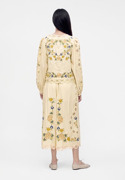 Femme aux longs cheveux foncés portant une tenue beige deux pièces avec broderies florales et détails en dentelle, debout de dos sur un fond blanc.