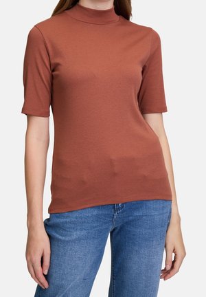 Kobieta ubrana w rdzawy, krótkim rękawem top z podwyższonym kołnierzem oraz niebieskie dżinsy, stojąca na tle gładkiej białej ściany.