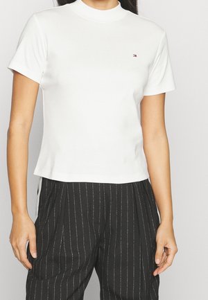 Femme portant un haut blanc à manches courtes ajusté avec col montant et un pantalon rayé noir taille haute.