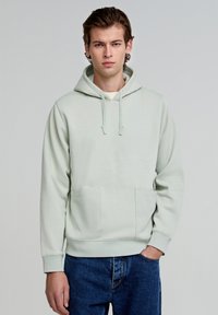 Lichtgroene hoodie van zachte stof, voorzien van een kangoeroezak, verstelbare trekkoordkap en ribgebreide manchetten en onderkant. Gecombineerd met donkerblauwe jeans.
