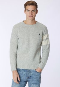 Maglione a maglia grigio chiaro con polsini e orlo a costine, caratterizzato da un emblema nero e righe crema su una manica. Indossato con jeans blu.