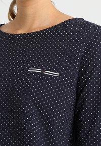 Navy blue long-sleeve shirt with white polka dots. Features a small chest pocket with a striped design and a red accent button.