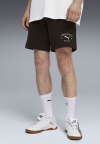 Shorts de sport en coton brun avec logo blanc, assortis à des chaussettes blanches et des chaussures de sport blanches avec des accents gris. Le fond passe du gris clair au gris foncé.