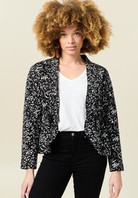 Blazer ajustado de patrón floral en blanco y negro, con mangas largas y dos bolsillos frontales, combinado con una blusa blanca de escote en forma de V.