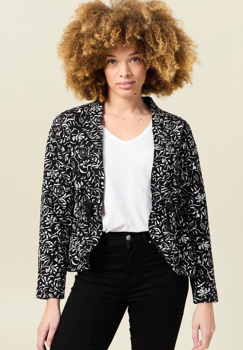 Blazer ajustado de patrón floral en blanco y negro, con mangas largas y dos bolsillos frontales, combinado con una blusa blanca de escote en forma de V.