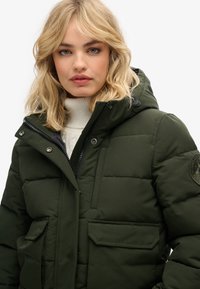 Chaqueta acolchada verde oscuro con cierre de cremallera y botones a presión, que cuenta con capucha, dos bolsillos frontales y un acabado de tela texturizada.