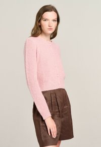 Rosa gestrickte Strickjacke mit rundem Ausschnitt, langen Ärmeln und Knopfverschluss, kombiniert mit braunen plissierten Shorts. Weiche Textur und taillierter Schnitt.