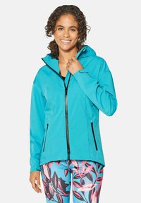 Türkise Softshell-Jacke mit Kapuze, durchgehendem Reißverschluss und schwarzen Reißverschlusstaschen. Kombiniert mit gemusterten Leggings mit pink- und schwarzfarbenen Blättern.