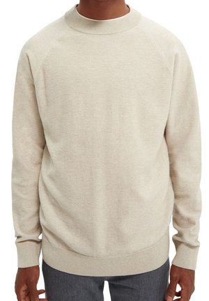 Maglione beige a maniche lunghe in maglia con polsini e collo a coste indossato da una persona abbinato a pantaloni grigio scuro.