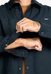 Mörkblå skjorta med knappar och textilstruktur. Har kontrasterande svarta manschetter, silverdetaljer och synliga tatueringar på händerna.