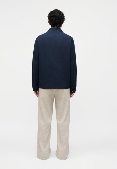 Άνδρας με σγουρά μαλλιά που φοράει μπλε σκούρο πουκάμισο με μακρύ μανίκι και μπεζ παντελόνι με φαρδιά πόδια, στέκεται με την πλάτη προς το μέρος μας σε απλό φόντο.