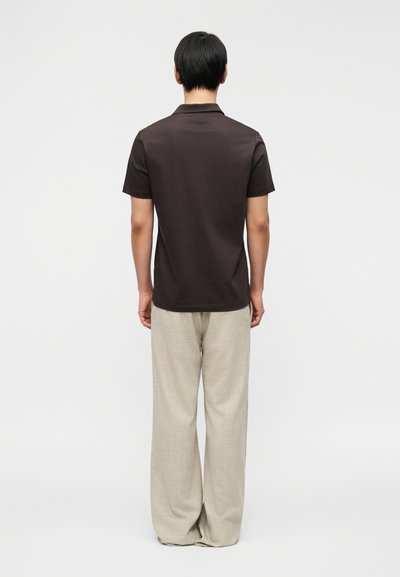 Καφέ κοντομάνικο πουκάμισο πόλο με γιακά, σε συνδυασμό με ανοιχτό μπεζ παντελόνι με φαρδιές φάσες. Απλός σχεδιασμός χωρίς ορατά σχέδια ή λεπτομέρειες.
