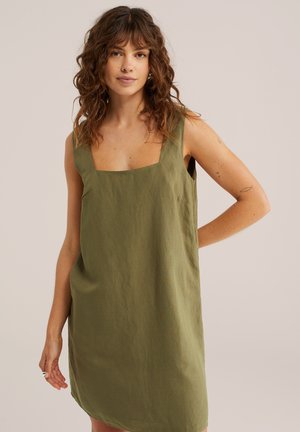 Žena s kučeravými hnedými vlasmi, oblečená v bezrukávové olivovo-zelenej šatách so štvorcovým výstrihom a tetovaniami na pravej ruke, stojaca pred jednoduchým pozadím.