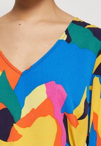 Blouse multicolore avec un col en V, ornée de formes abstraites audacieuses en bleu, orange, rose, vert et jaune sur un tissu lisse.