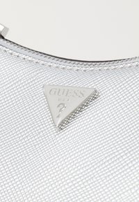 Borsa a tracolla argentata con texture, dotata di una targa triangolare in metallo che riporta la scritta "GUESS U.S.A." con un'icona di punto interrogativo. Chiusura con cerniera.