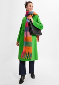 Green wool coat with large lapels, black handbag, colorful striped scarf in orange, pink, and blue, dark flared jeans, and black heeled boots.
