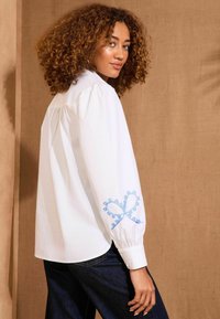 Camicia bianca a maniche lunghe con maniche a sbuffo, con dettagli ricamati blu sui polsini. Il tessuto ha una texture liscia.