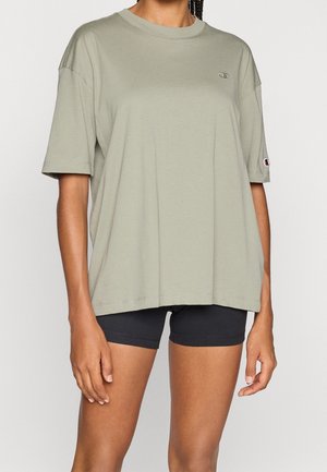 Osoba nosząca oversize jasnoszary t-shirt z krótkim rękawem i czarne dopasowane krótkie spodenki rowerowe, stojąca z rozluźnionymi ramionami.