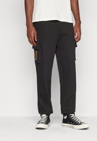 Schwarze Cargo-Sweatpants mit Seitentaschen, mit einem gestickten goldenen Design auf der linken Tasche; kombiniert mit schwarzen High-Top-Sneakern.