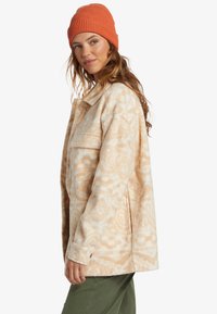 Fleece jack in licht crème met abstracte tan patronen; heeft een kraag en een voorkantoor. Gecombineerd met een oranje gebreide muts.