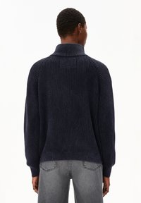 Eine Person mit kurzem, lockigem Haar trägt einen dunkelblauen, gerippten Strickpullover und graue Denim-Jeans. Sie steht mit dem Rücken zu einem schlichten Hintergrund.