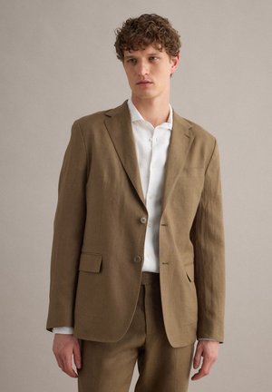 Joven vestido con traje marrón claro y camisa blanca, de pie frente a un fondo neutro y sencillo, mirando ligeramente de lado.