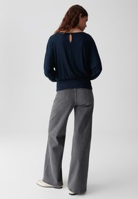 Navy-Blaues Stricktop mit lockerer Passform, das ein Schlüsselloch-Detail am Rücken aufweist; kombiniert mit grauen Schlagjeans und hellen Schuhen.