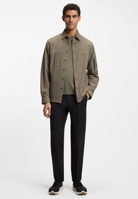 Koszula khaki na guziki z kołnierzykiem i dwoma kieszeniami na piersi, zestawiona z zielonym T-shirtem i czarnymi spodniami, uzupełniona czarnymi trampkami.