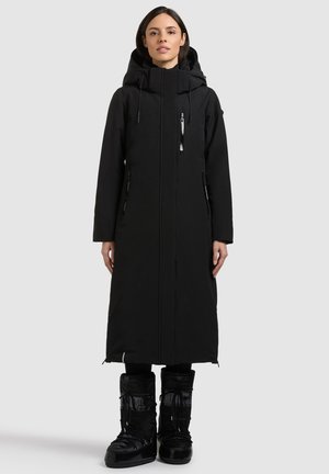 Frau steht, trägt einen langen schwarzen Wintermantel mit Kapuze und schwarze isolierte Winterstiefel vor einem schlichten hellen Hintergrund.