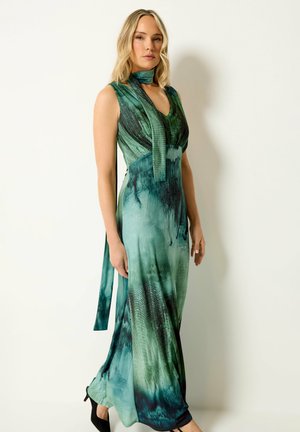 Mujer con vestido maxi sin mangas de tie-dye verde y turquesa, con pañuelo a juego en el cuello y zapatos negros de tacón, de pie frente a una pared lisa.