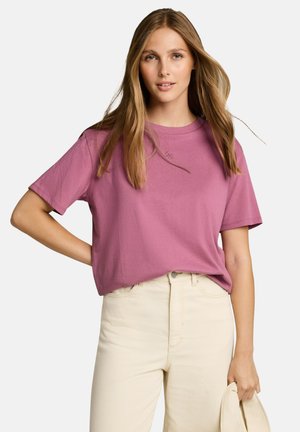 Junge Frau mit langen blonden Haaren trägt ein rosa kurzärmeliges Shirt und cremefarbene Hose mit hoher Taille und hält eine passende cremefarbene Jacke in der Hand.
