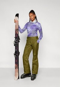 Smučarska obleka s vijoličnim in belim vzorčastim dolgim rokavom, zelenimi hlačami in črnimi smučarskimi čevlji, ki drži črno smučko z vezmi.