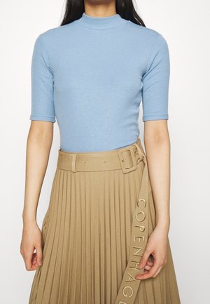 Vrouw draagt een lichtblauw, geribbeld T-shirt met korte mouwen, ingestopt in een beige geplooide rok met een bijpassende riem en een gesp met de tekst "COPENHAGEN" erop geborduurd.