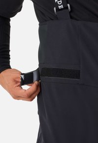 Schwarze wasserdichte Latzhose mit verstellbaren Trägern und einer strukturierten Klettverschlusslasche an der Seite. Das Material scheint strapazierfähig und flexibel zu sein.