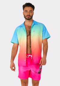 Short-sleeve shirt with gradient blue to pink color, button closure, paired with bright pink shorts, holding sunglasses.