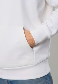 Inimene, kes kannab valget pusat käega ees taskus, nähtavad õmblused ja sinised teksad neutraalse tausta ees.