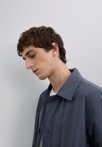 Joven con cabello castaño que lleva un abrigo azul, mirando hacia abajo pensativamente contra un fondo gris claro liso.