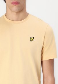 Short-sleeved, round-neck t-shirt in light yellow cotton. Features a black-outlined yellow bird logo on the left chest. Soft texture.