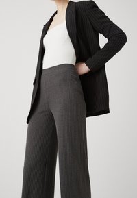 Woman wearing a black pinstripe blazer over a white ribbed top and high-waisted gray ribbed wide-leg pants, standing with hand on hip.