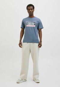 Blauwe katoenen t-shirt met een zwart en roze "JACK&JONES" graphic, gecombineerd met crèmekleurige wijde broeken en grijze sneakers, staande tegen een plain achtergrond.