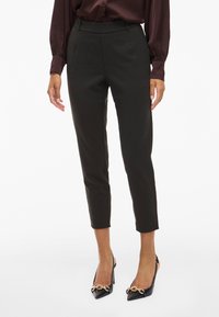 Črne krojene hlače z visokim pasom, skrajšano dolžino in gladkim materialom. V kombinaciji s črnimi salonarji z ostrim nosom, ki imajo zlat detajl verige.