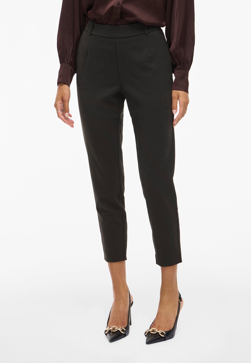 Črne krojene hlače z visokim pasom, skrajšano dolžino in gladkim materialom. V kombinaciji s črnimi salonarji z ostrim nosom, ki imajo zlat detajl verige.