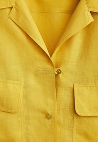 Veste en lin jaune avec col cranté, poches plaquées à l'avant et deux boutons visibles pour la fermeture.