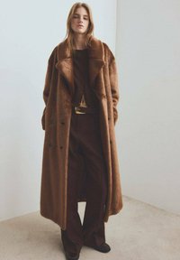 Brauner Pelzmantel aus Kunstfell mit weitem Kragen, Knopfverschluss und Seitentaschen, über einem dunkelbraunen Outfit und braunen Schuhen getragen.