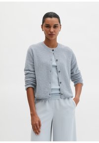 Hellblauer, strukturierter Cardigan mit rundem Ausschnitt, Knöpfen vorne und gerafften Ärmeln, kombiniert mit einem passenden, gerippten Top und hellen Hosen.