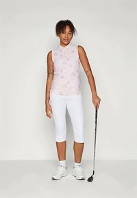 Sieviete bez piedurknēm ar ziedu raksta polo krekls un baltas capri bikses, tur rokās golfa nūju, stāvot pret vienkāršu neitrālu fonu.